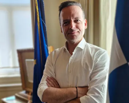 Haris Doukas standing between the Greek and European Union flags in his office.