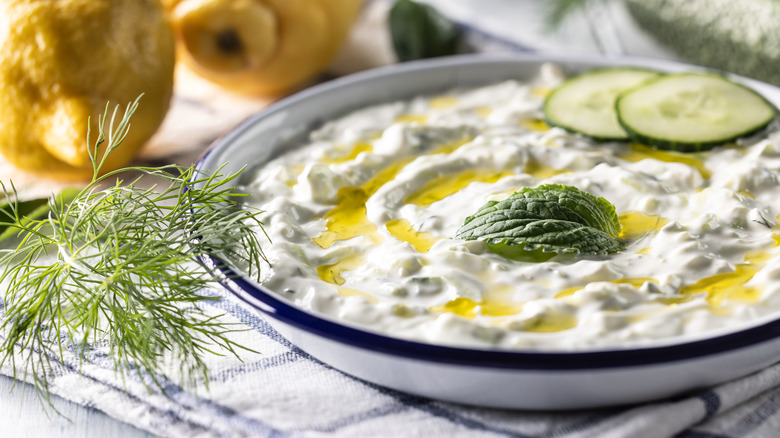 Arka planda limonlar olan, salatalık dilimleri, nane ve dereotu ile süslenmiş bir kase cacık