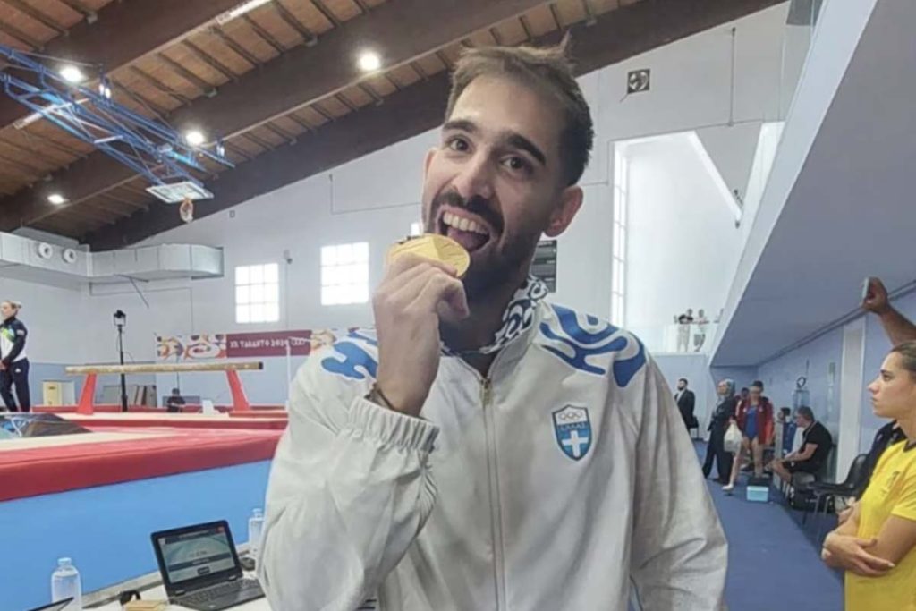 Sporcu kapalı spor salonunda altın madalyasını ısırıyor, arma beyaz ceket giymiş, kutlama pozu.